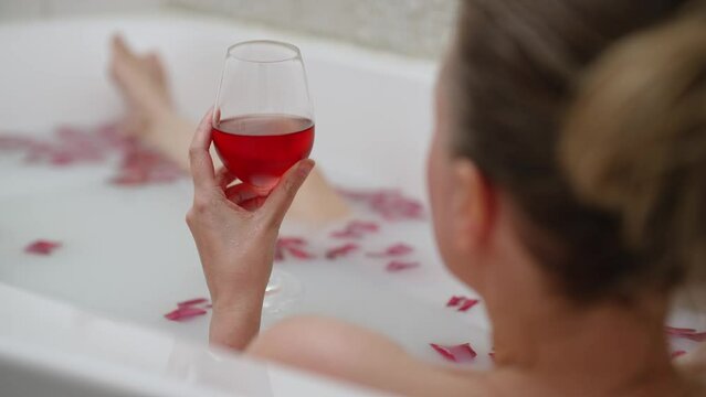 Woman enjoys wine in a bath with milk and rose petals.