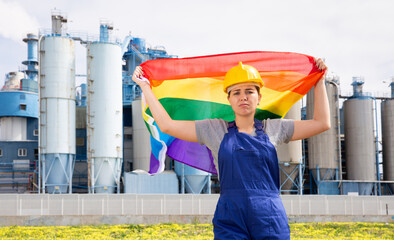 Sad female worker in hardhat with rainbow flag standing in front of factory