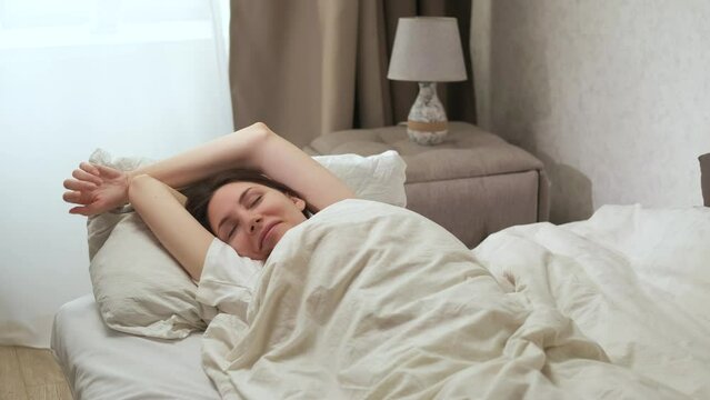 A lazy pretty young woman stretches in bed, smiles, opens her eyes in the morning and wakes up