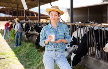 Confident asian woman dairy farm owner standing with glass of fresh milk near stalls with cows on sunny summer day