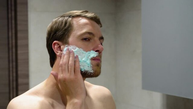 Shirtless Man Applying Shave Foam On Arm While Standind Near Mirror In Bathroom, Smiling Guy Preparing For Shaving, Making Morning Beauty Routine At Home