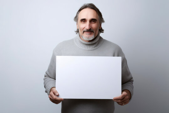 Handsome middle-aged man in a gray sweater holding a white sheet of paper on a gray background