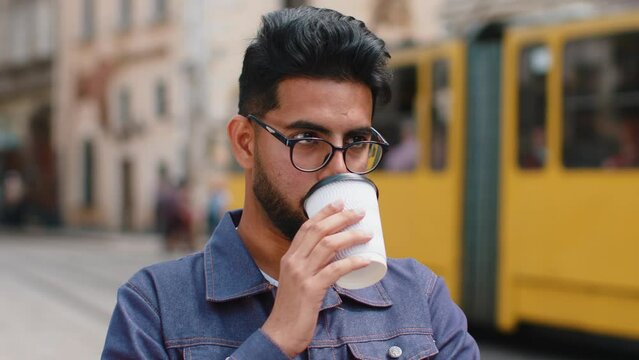 Young indian man in glasses enjoying morning coffee or tea hot drink outdoors. Relaxing, taking a break. Hindu guy walking in urban city center street, drinking coffee to go. Town lifestyles outside