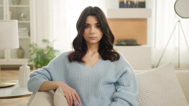Beautiful Latin American woman in blue sweater looking at camera sitting on sofa alone at home. Calm pleasant young woman feels relaxed posing portrait in living room of bright modern design apartment