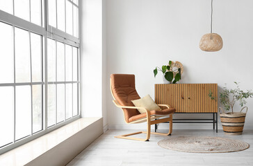 Cozy brown armchair with cushions and wooden cabinet near big window in interior of living room