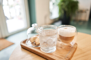 Cup of coffee cappuccino with cantuccini and a glass of water on a wooden tray. Coffee with milk, a cookie and a glass with water on a glass table.