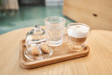 Cup of coffee cappuccino with cantuccini and a glass of water on a wooden tray. Coffee with milk, a cookie and a glass with water on a glass table.