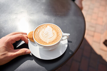 A cup of hot cappuccino with cookies on the table in the coworking space of the coffee shop. A cup of coffee on the veranda on a sunny day. Coffee break at the cafe. Close-up