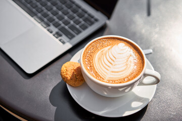 A cup of coffee in hand next to laptop. A cup of hot cappuccino coffee with cookies and a laptop computer on the table in the coworking space of the coffee shop. Online business, work in cafe