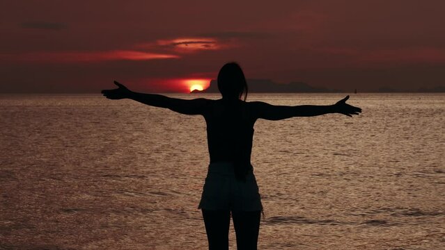 Silhouette of woman looks at sun disk on sky during red sunset on the sea, before sun goes below horizon. Woman greets sunset with her arms outstretched and enjoying beautiful seascape while traveling