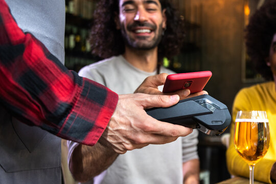 Unrecognizable male costumer paying round of beers in a bar with mobile phone. Waiter holding dataphone. Focus on hands.