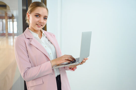 image of a successful business woman female employee of a company Smiling and looking at camera holding laptop standing by mirror in office
