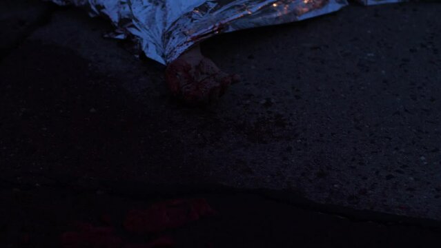 A anonym dead person killed in a car accident. Covered by a silver foil. Bloody hand is showing up. Flashing sirens in the back.