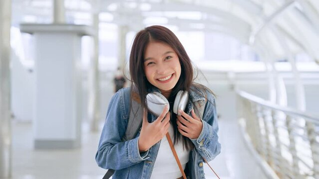 Young asian woman wearing denim jacket, headphones on her neck and backpack walking in the city. Adolescent female happy, smiling face. Young lady relaxing and enjoying city life