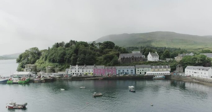 Wide drone shot panning across a bay in Loch Portree showing the colourful houses on the shore, along with boats moored in the bay in Portree on the Isle of Skye