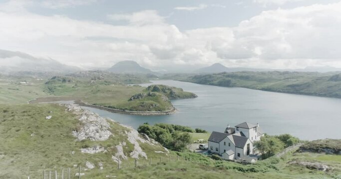Slow drone panning shot of a mansion house in the foreground, a Loch in Scotland in the middle and tall mountains and clouds in the distance and sun shining