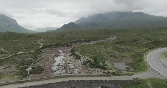 Slow drone reveal along a river revealing the Sligachan Old Bridge on the Isle of Skye with traffic crossing a bridge on the river