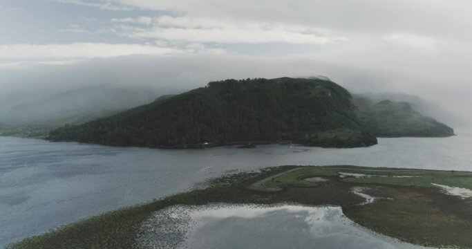 Wide drone shot of a cloud covered coastal mountain with the mist and cloud rolling over the sea and mountain peak in Scotland