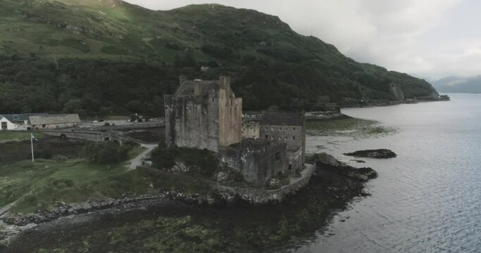 Close up drone shot of Eilean Donan Castle moving backwards to reveal it is next to an estuary and surrounded by mountains in Scotland