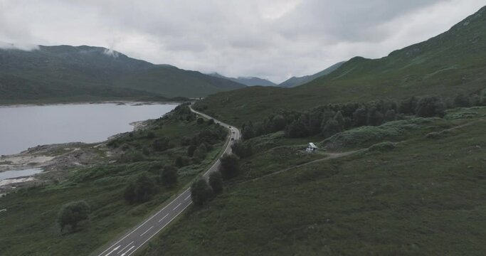 Wide push in drone shot of Loch Beag, traffic travelling along the coastal road and a campervan wild camping with BBQ smoke and mountains in the background in the Scottish Highlands