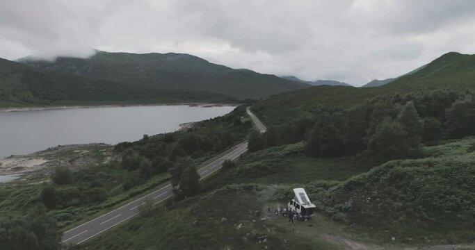 Static mid drone shot showing a campervan wild camping with BBQ smoke and Loch Beag and cloudy mountains in the background in Scottish Highlands