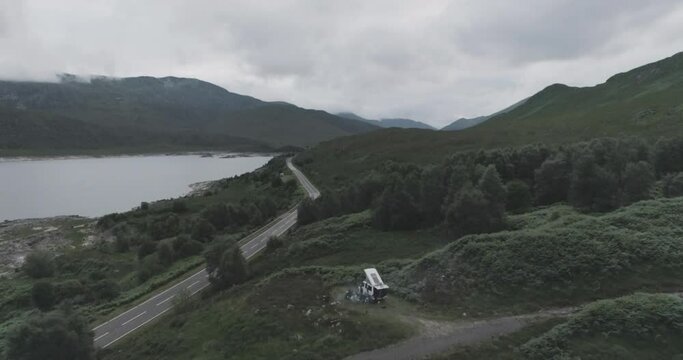 Close drone shot showing a campervan wild camping with BBQ smoke and Loch Beag and cloudy mountains in the background in Scottish Highlands