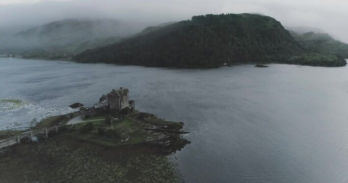 Slow wide drone reveal of Eilean Donan Castle, mist and wind over the sea and cloud covered mountains in the background in Scotland