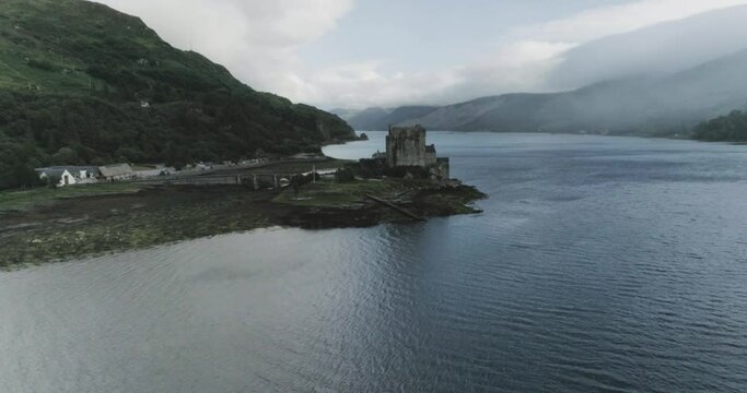 Mid drone shot showing Eilean Donan Castle alongside the sea and scottish flag flying with mist and cloud over the water in Scotland