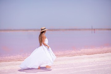Woman in pink salt lake. She in a white dress and hat enjoys the scenic view of a pink salt lake as she walks along the white, salty shore, creating a lasting memory.