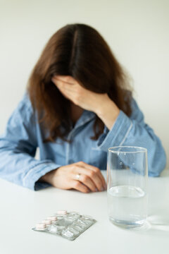 A young girl in pajamas sits at a table and holds her head against the background of a glass of water with pills. The concept of PMS, migraine, stress, depression.
