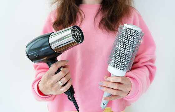 The girl holds a hair dryer and a brush in her hands. Drying long brown hair with a round brush for styling unruly hair. Beauty saloon.