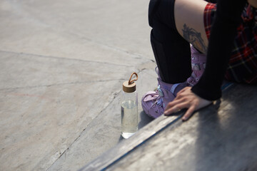 Skater girl sitting on a ledge with a glass bottle of fresh water. Roller blader female wearing modern aggressive inline skates in a skatepark and chilling after the ride