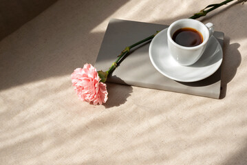 Minimalist aesthetic summer still life, coffee cup, notebook and carnation flower on beige table with abstract lifestyle sunlight shadows