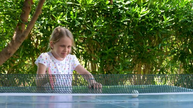 Smile girl play table tennis, happy childhood. Child player hit lightweight ping-pong ball back and forth across hard table divided by tennis net use small red rackets. Sport training in garden