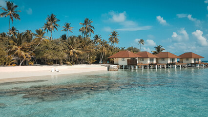 Palafitte in villaggio maldiviano con spiaggia bianca, mare cristallino, palme  e coppia di sdraio...