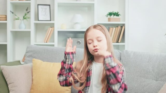 Charming little girl in checkered shirt resting alone on comfy couch and moving rhythmically with hands. Pretty caucasian kid enjoying favorite music with closed eyes during free time at apartment.