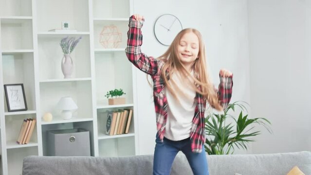 Cheerful little lady feeling happy and jumping joyfully on soft couch. Casually dressed girl fooling around and enjoying school holidays at home. Concept of leisure activity and positive emotions.
