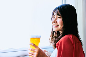 明るい窓際でグラスに入ったビールを持って笑顔で味わいながら遠くを眺める30代の1人の女性