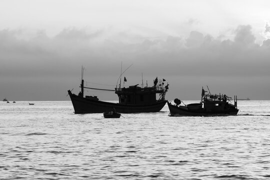 Black and white photo at sea with silhouettes of fishing boats and Vietnamese fishermen in the morning. The image has a high contrast of black and white.