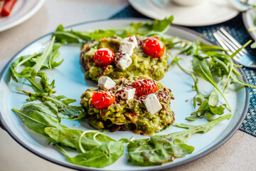 Healthy breakfast with avocado toast, greek cheese, and cherry tomatoes on rye bread. A delicious vegetarian meal in a green outdoor cafe, plant-based clean food and a healthy fat option.