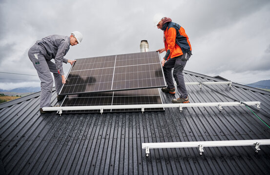 Technicians installing solar panel system on roof of house. Men Installers in helmets carrying photovoltaic solar module outdoors. Concept of alternative and renewable energy.