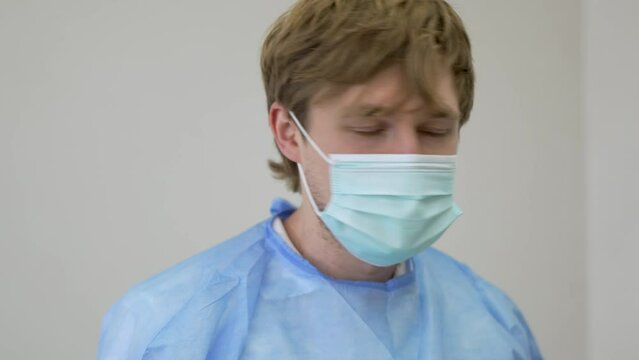 Front view of young blond male dentist wearing a mask and medical uniform turning his head and looking for a while at the viewer