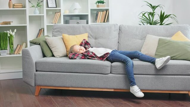 Exhausted preteen child taking nap on comfortable couch after intense working day at school. Cute little girl regaining strength and sleeping in well-decorated trendy apartment.