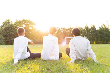 Three boys are sitting with their backs to us on the soccer field in white shirts at sunset
