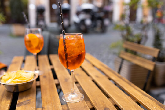 Aperol Spritz Cocktail. Alcoholic beverage based on table with ice cubes and oranges outdoors. Served cocktail with orange slice and straw placed on wooden table of sidewalk cafe in Italy