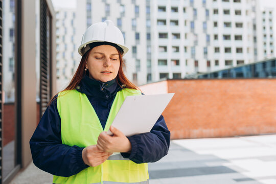 Architect with paper folder with documents at a construction site. Woman constructor wearing helmet and safety yellow vest. Women are planning new building project. Modern exterior