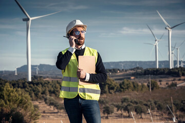 Mature male engineer talking on a walkie talkie with his colleague while checking and taking notes on the operation of wind turbines - obrazy, fototapety, plakaty