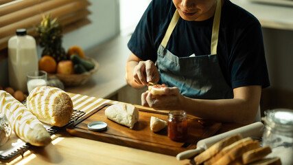 Man in apron spreading raspberry jam on slice of bread with table knife, preparing a healthy breakfast at kitchen table