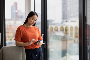 Young asian businesswoman in corporate office with view of city skyline using smartphone