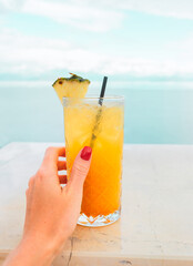 Female hand with a red manicure holds a tropical yellow-orange cocktail on the background of the sea. Vacation photo